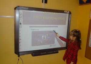 Dziewczynka stoi pod tablicą interaktywną i wskazuje symbole Unii Europejskiej.